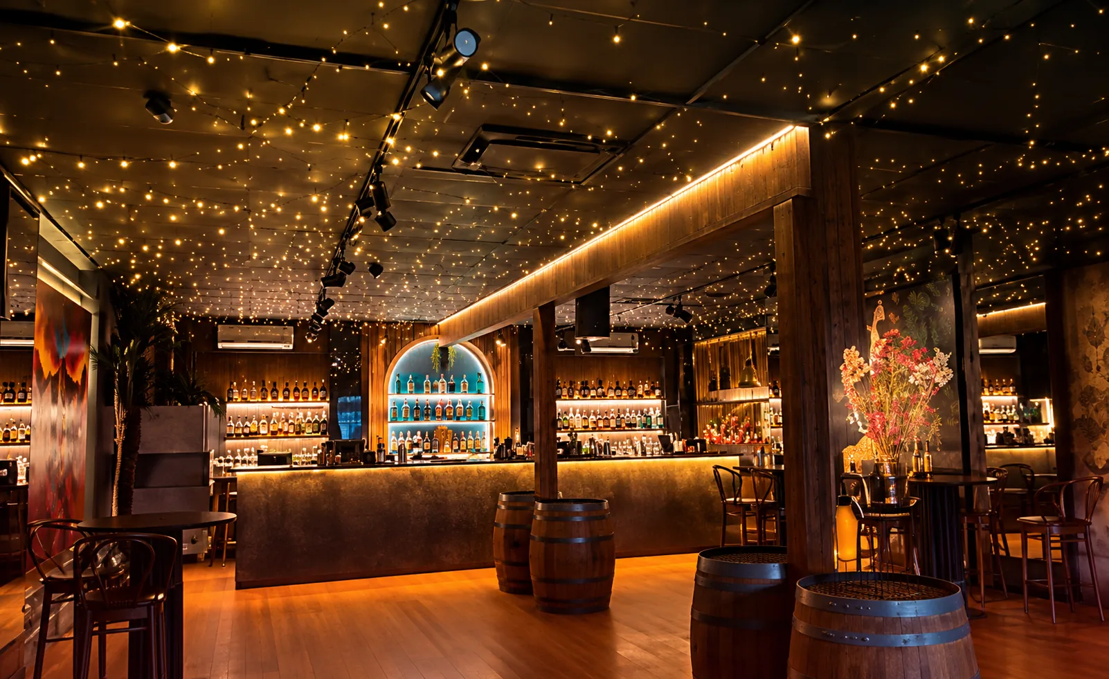 Trombar at night: barrels and high tables beneath a ceiling of warm fairy lights, with the backlit bar and a blue-lit arch at the far end